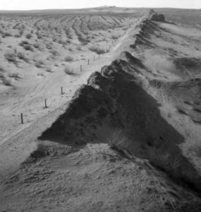 The landscape is sandy dunes. Poking out of the sand like spines are broken remnants of a wall. This image demonstrates the erosion caused by desertification. 