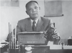 Photograph of Ho Fengshan a middle aged man wearing a business suit sitting at a desk in front of a typewriter. 