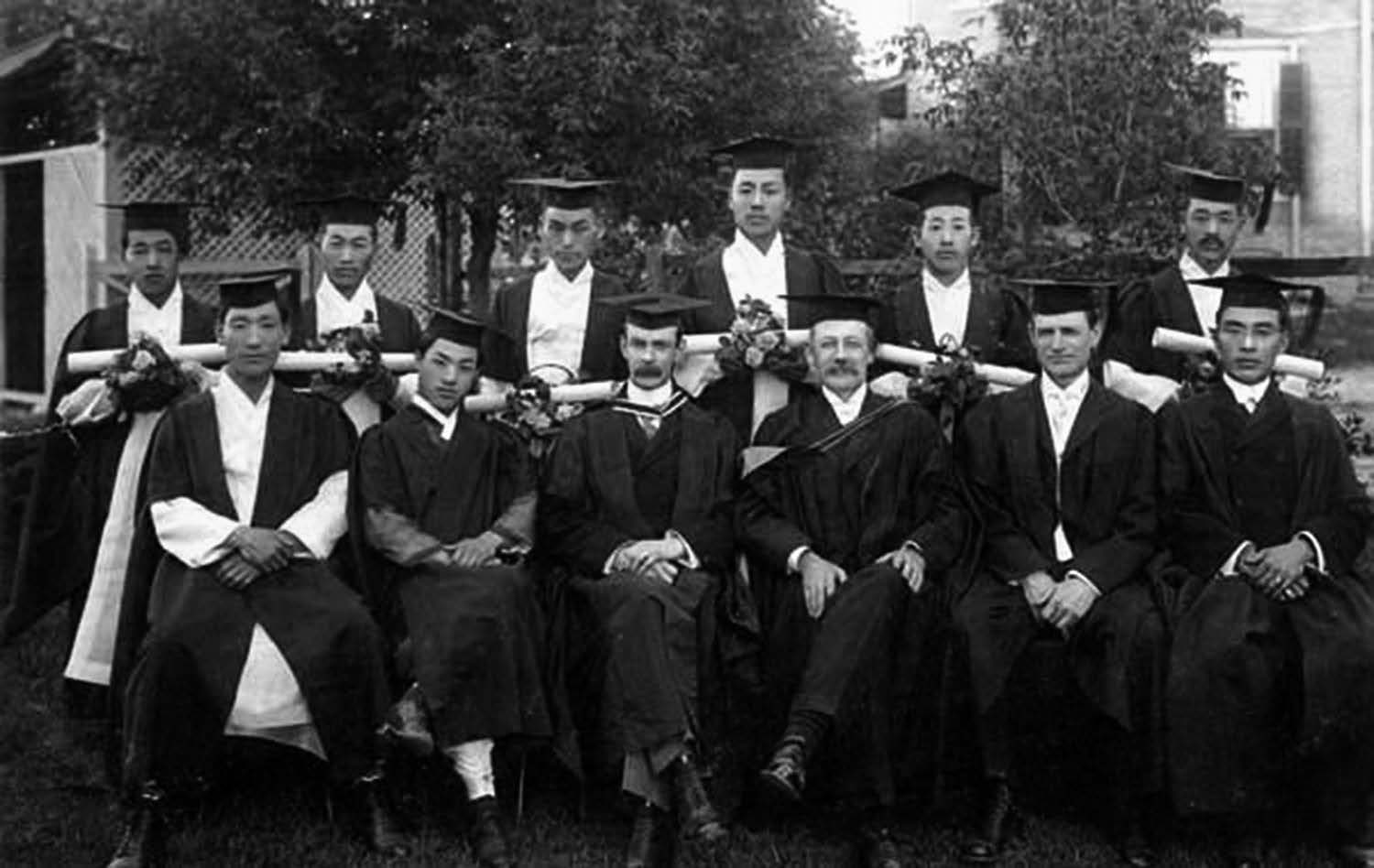 1911 Severance Union Medical College graduates (six graduates in total) are shown with members of the faculty: Dr. Kim, Dr. Hirst, Dr. Avison, Dr. Weir, Dr. Pak, Dr. Hong. The students and faculty are all wearing graduation robes. 