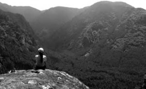 Photo of the backside of a woman sitting on top of a mountain staring out into the mountainous landscape. 