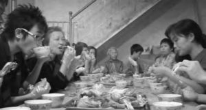 A group of people sit at a long table while eating chicken. 