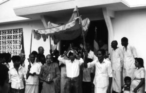 many people in attendance face a man holding something walking toward the camera. People hold a tent over his head and the relic