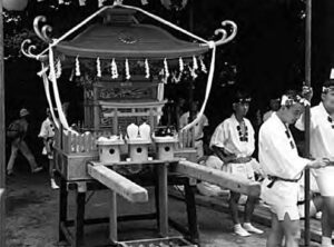 a photograph of a shine palanquin, with several men in white robes stand nearby. At the front of the palanquin is two long wooden rods, where the people would hold it when they are picking it up.