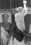 a white statue in a sea of protest flags looks at a distant portrait of a man