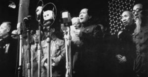 a photo of a man reading from a paper and speaking into several microphones. several old and middle aged men stand behind him