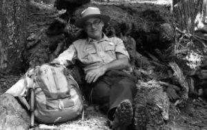 A man in glasses and a ranger's uniform lounges against a pile of dirt and rocks. His hiking backpack sits next to him.