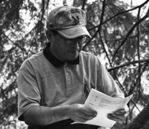 a man in glasses and a hat looks down at a packet while outside underneath tree branches.