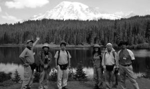 Six people stand in front of a lake and a nearby mount fuji. One man proudly gestures towards the mountain while the rest face forward.