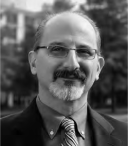 Photograph of Constantine Vaporis smiling at the camera. He is a middle aged man with a balding head and a greying beard. He is wearing glasses and a suit jacket.