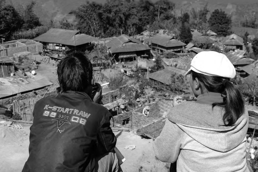 A woman and and man sit on a cliff looking down at their village. 