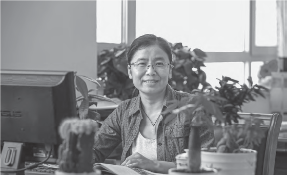 Wang Ping, a smiling middle-aged Asian woman wearing glasses, seated in front of a desk. The desk is equipped with a computer and adorned with a bamboo plant, creating a harmonious and productive work setting.